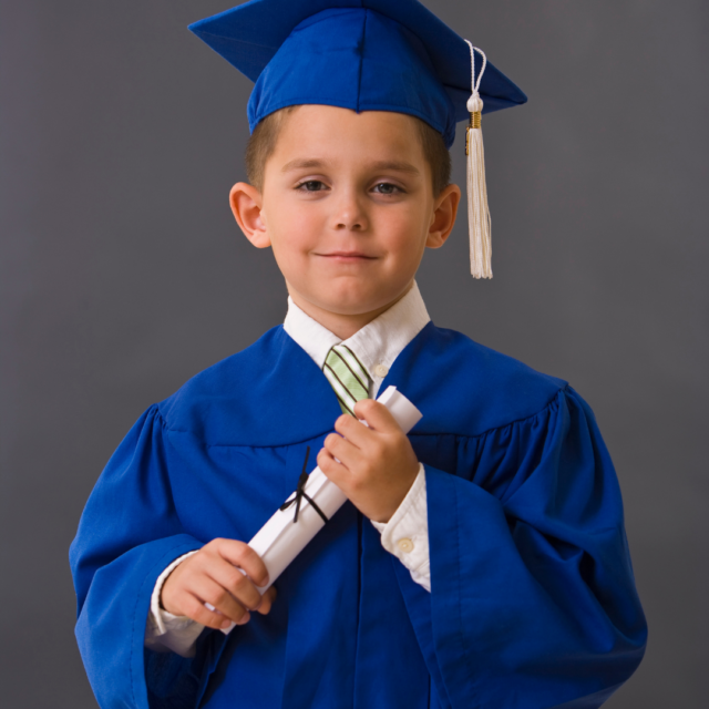 Young boy in a blue graduation cap and gown holding a rolled diploma tied with a ribbon in front of a gray backdrop, smiling for a graduation portrait.