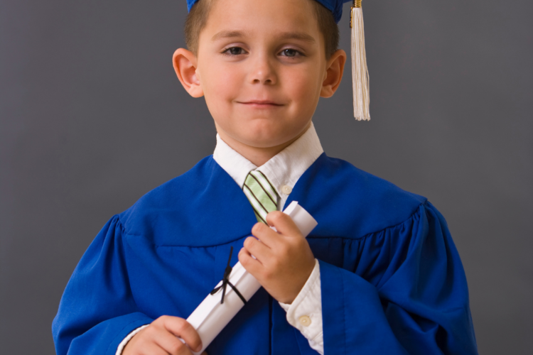 Young boy in a blue graduation cap and gown holding a rolled diploma tied with a ribbon in front of a gray backdrop, smiling for a graduation portrait.