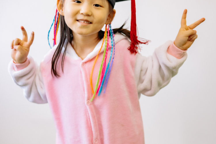 Child in a pink onesie wearing a graduation cap with colorful tassels, smiling and making peace signs with both hands and forearms raised.
