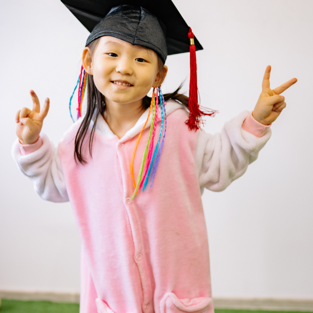 Child in a pink onesie wearing a graduation cap with colorful tassels, smiling and making peace signs with both hands and forearms raised.