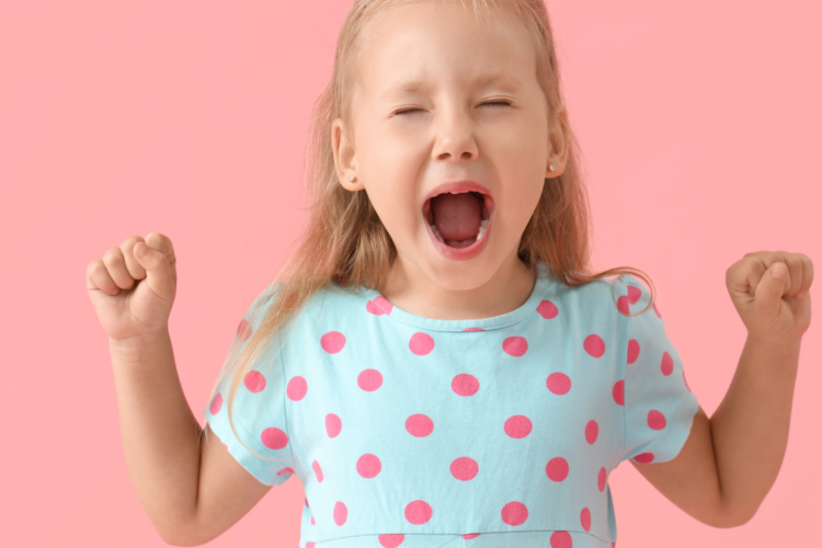 Little girl in a blue polka-dot dress raises fists and shouts, celebrating against a pink background.