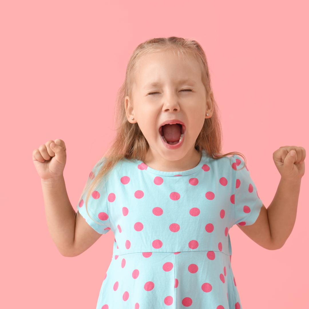 Little girl in a blue polka-dot dress raises fists and shouts, celebrating against a pink background.