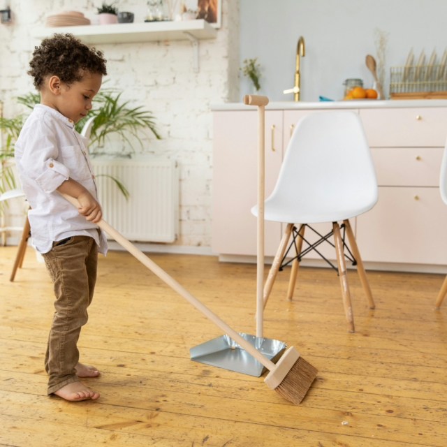 Young child sweeping with a broom into a dustpan in a bright kitchen.