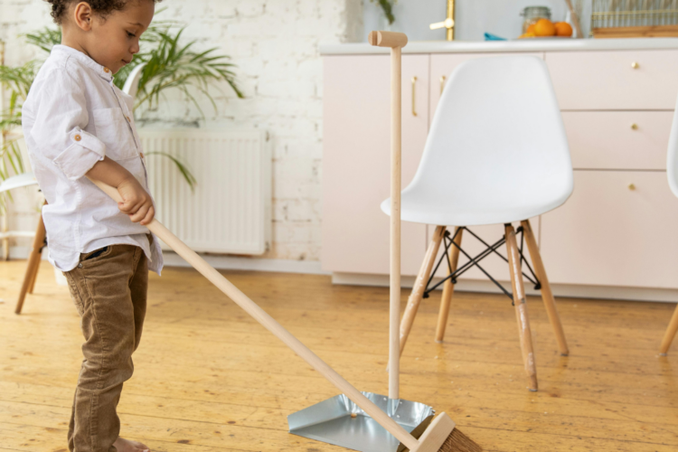 Young child sweeping with a broom into a dustpan in a bright kitchen.