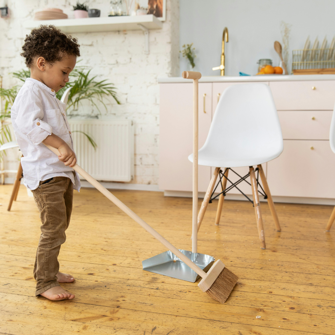 Young child sweeping with a broom into a dustpan in a bright kitchen.