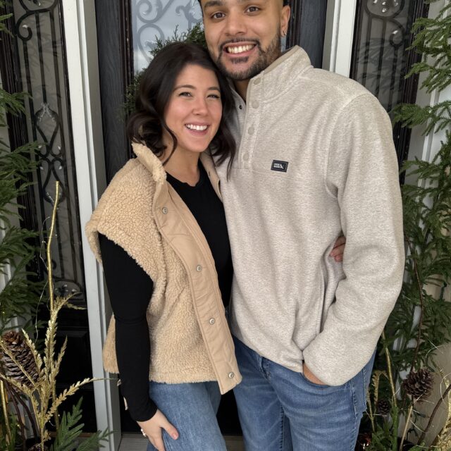 Smiling couple standing close together in front of a black door framed by greenery and holiday decorations