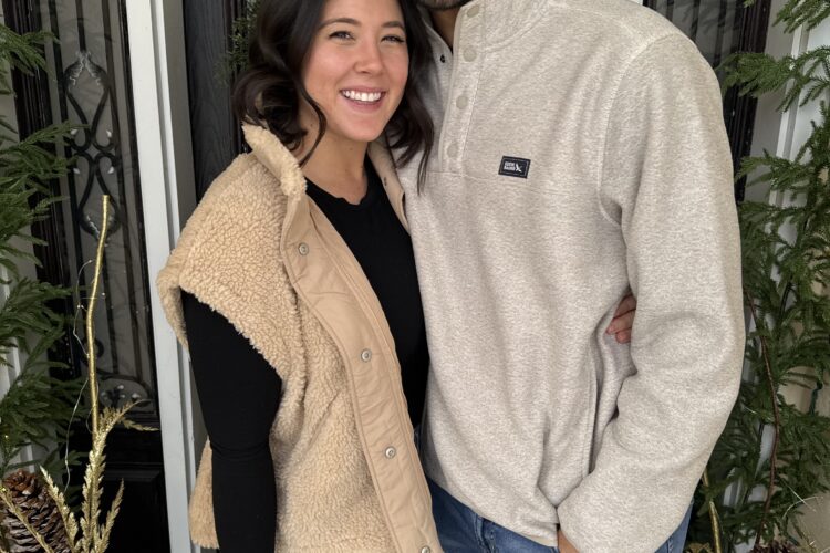 Smiling couple standing close together in front of a black door framed by greenery and holiday decorations