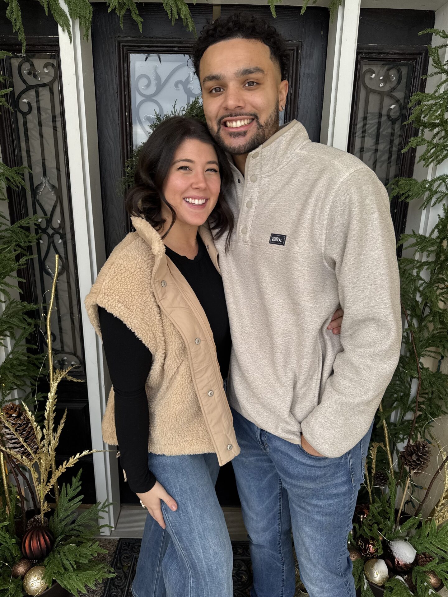 Smiling couple standing close together in front of a black door framed by greenery and holiday decorations