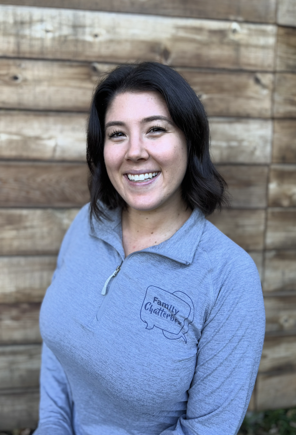 Smiling woman wearing a light blue zip-up top with a 'Family Clatterbox' logo, standing in front of a wooden fence.